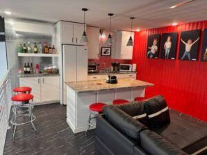 Modern storage display of kitchen and living room with red accents, island, black sofa, bar stools, and wall art
