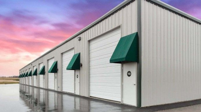 White storage building with green awnings and roll-up doors on a wet lot at sunset
