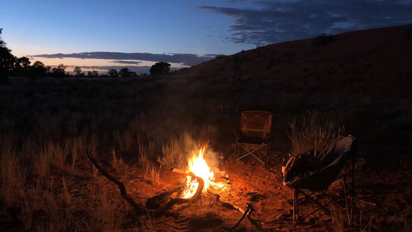 Campfire burning at dusk with a chair in a grassy field and a dark hill in the background.