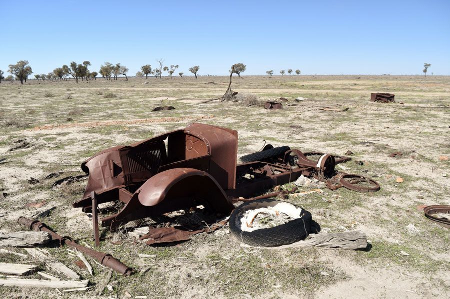 Rusty car wreckage in a dry, open field under a blue sky, surrounded by sparse vegetation.