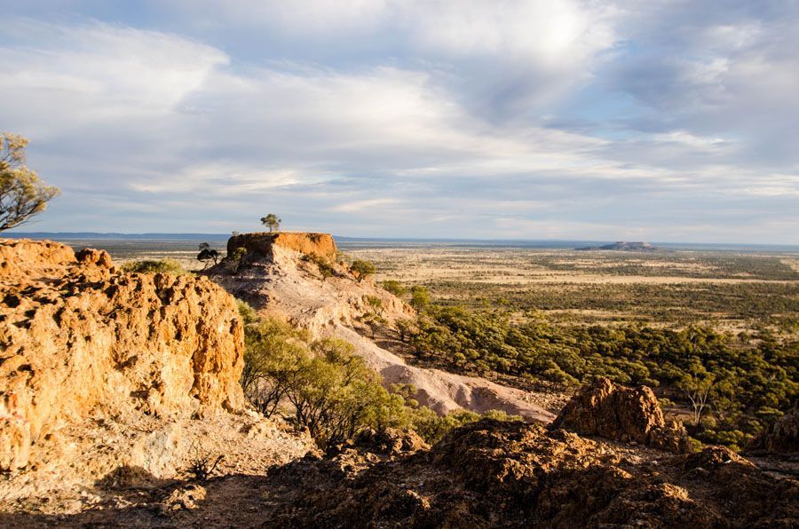 View of a desert landscape from a high, eroded cliff; golden light, cloudy sky, sparse vegetation.