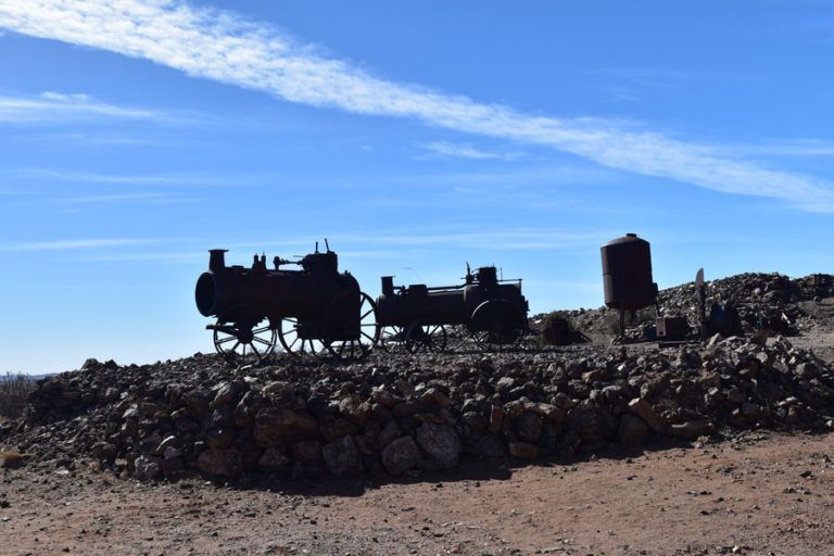 Silhouette of two antique steam engines and a water tank against a blue sky, on a rocky terrain.