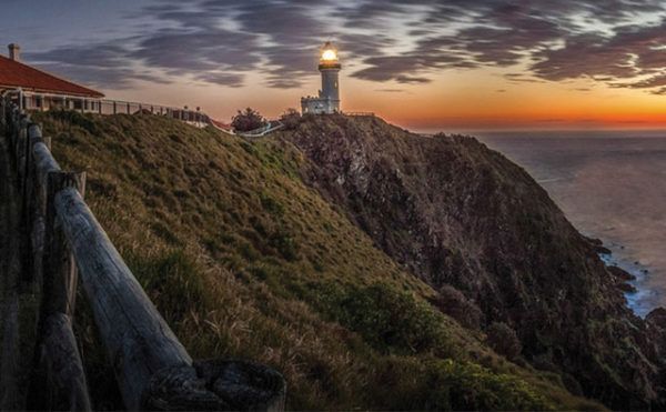 Lighthouse atop a cliff at sunset; ocean waves below, wooden fence in foreground; warm, orange, and blue hues.
