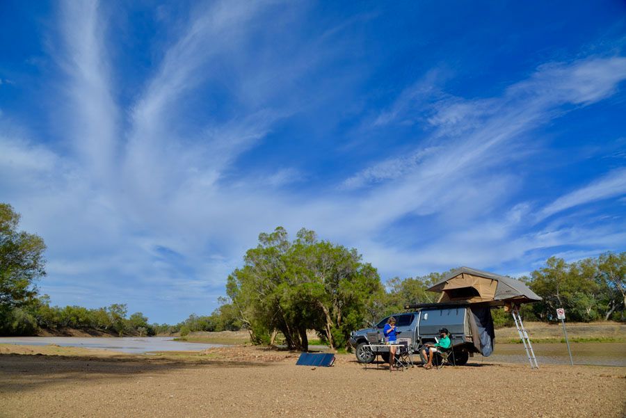 A 4x4 truck with a rooftop tent is parked near a riverbank under a blue sky with streaky clouds.