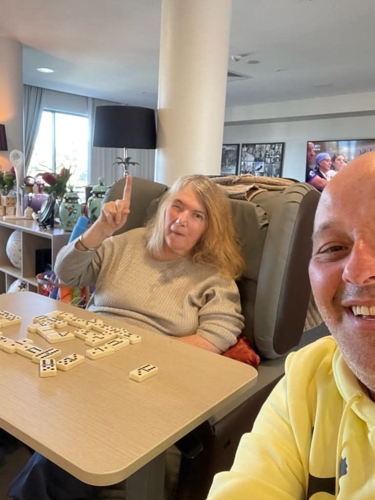 Woman pointing at dominoes while playing with a man. They are indoors at a table, smiling.
