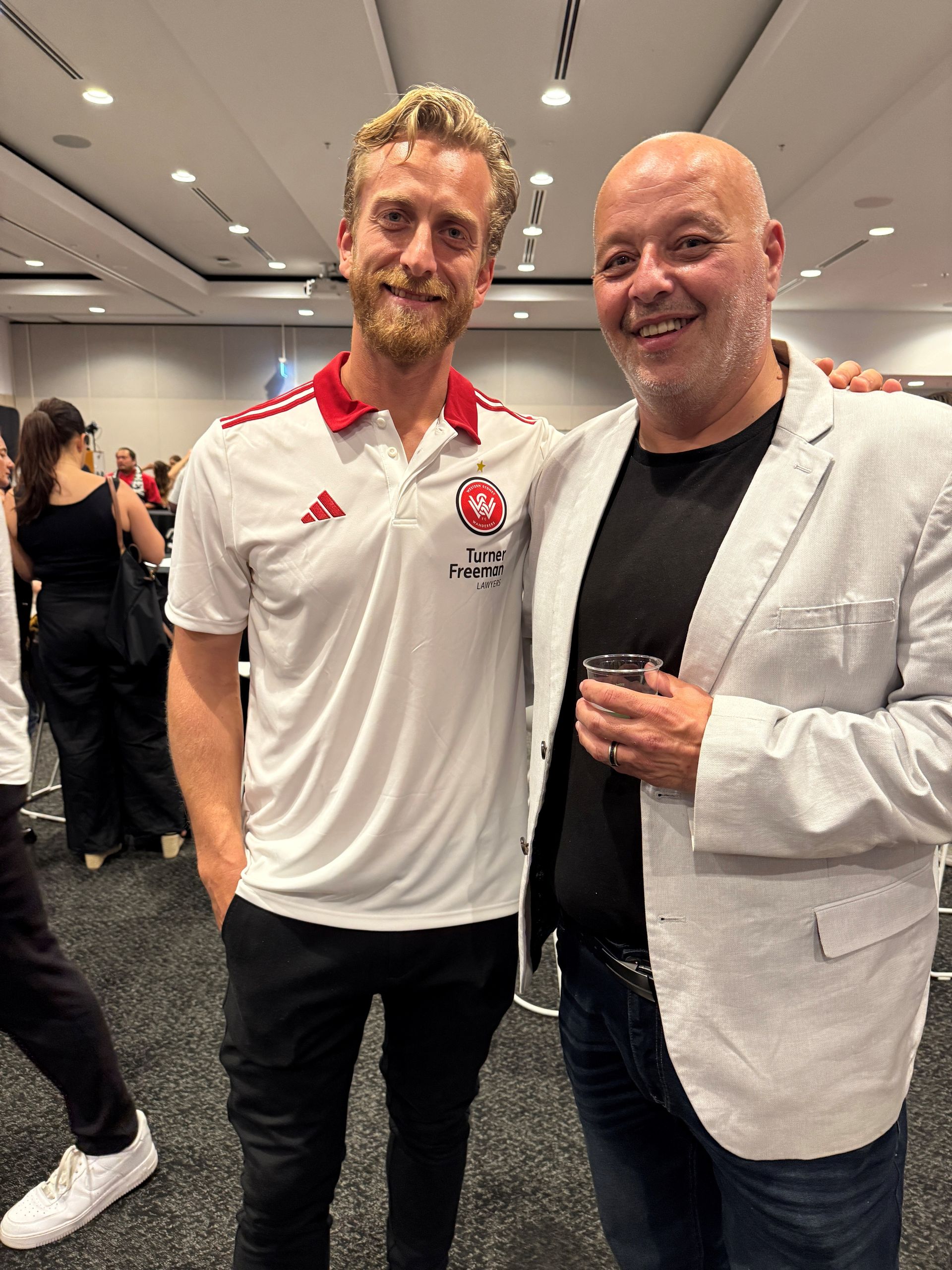 Two men smiling, posing. One in a white Chicago Fire FC polo, black pants. Other wears a blazer over a black top, jeans.