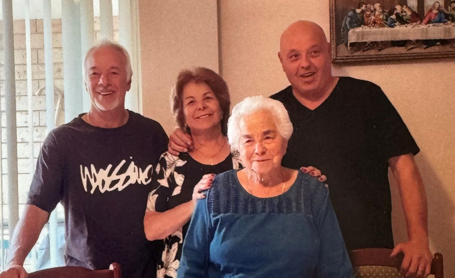 Four people, an elderly woman in blue shirt, smiling, with three others in a room with a framed artwork.