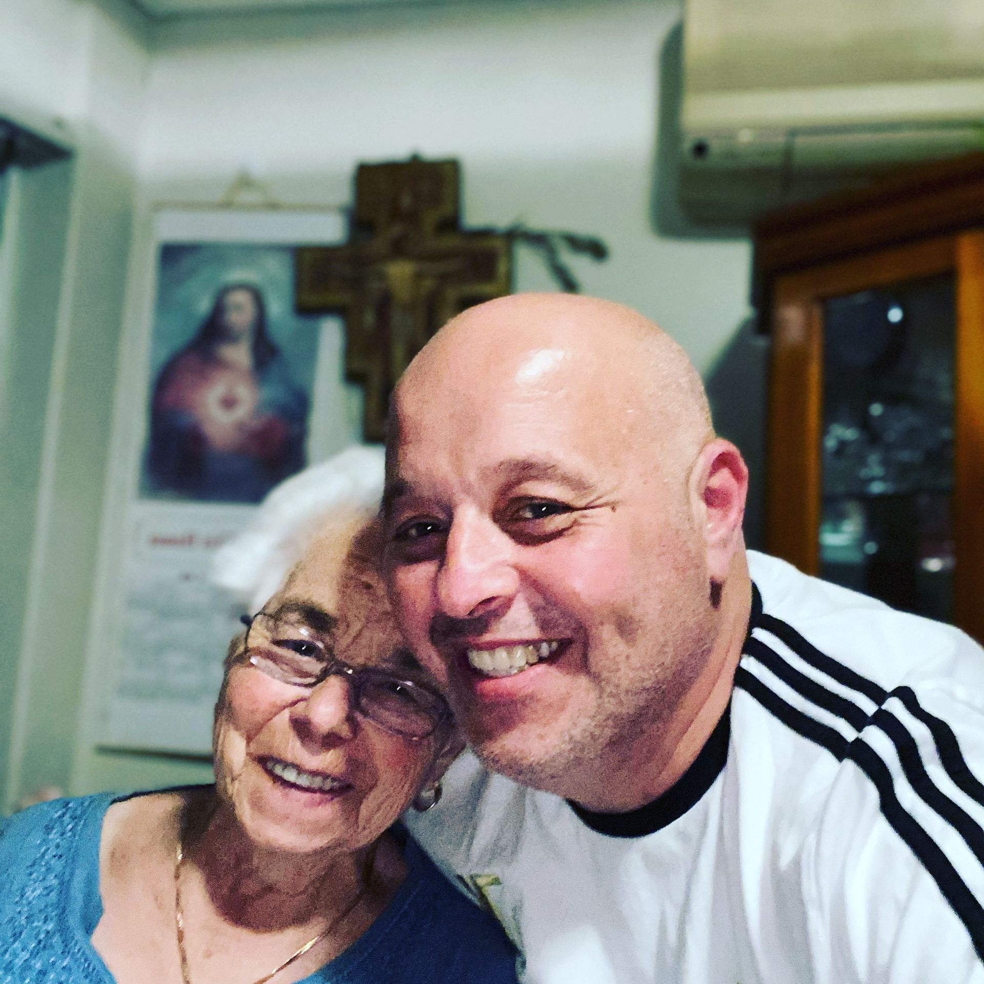 Smiling man with bald head and elderly woman, posing for selfie. Religious art in background.