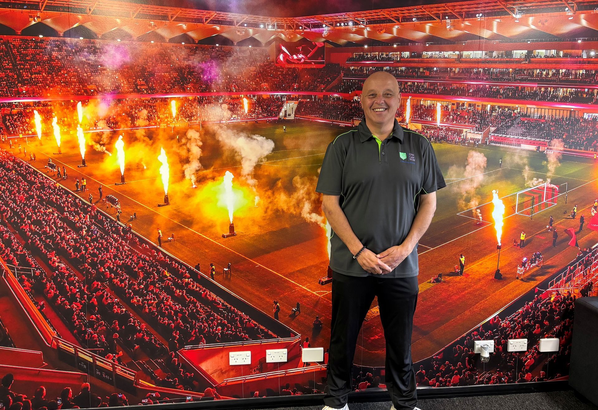 Man standing in front of a stadium mural with flames. He smiles, wearing a green shirt and black pants.