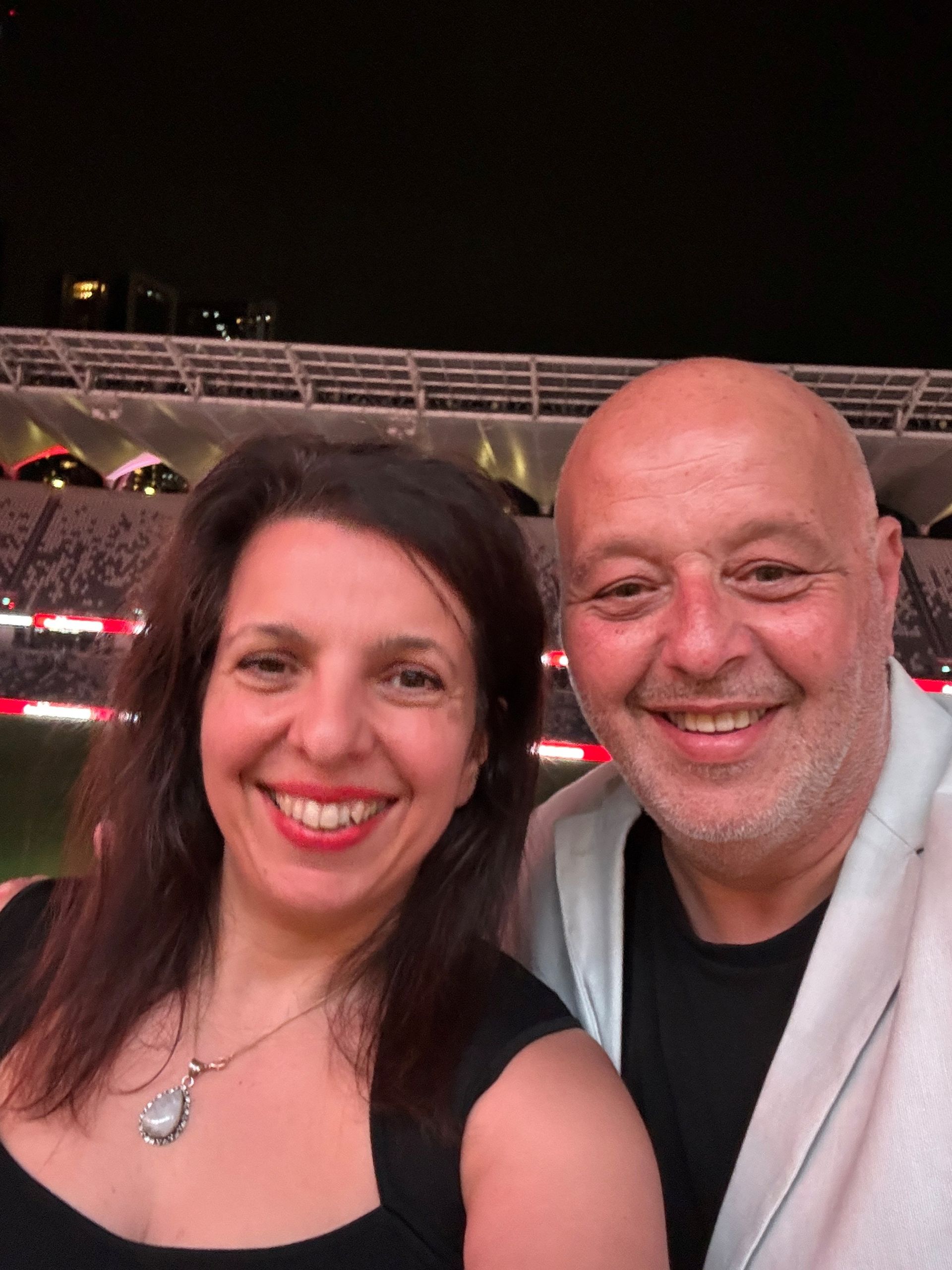 Smiling couple, woman with dark hair and red lipstick, man with bald head, at stadium.