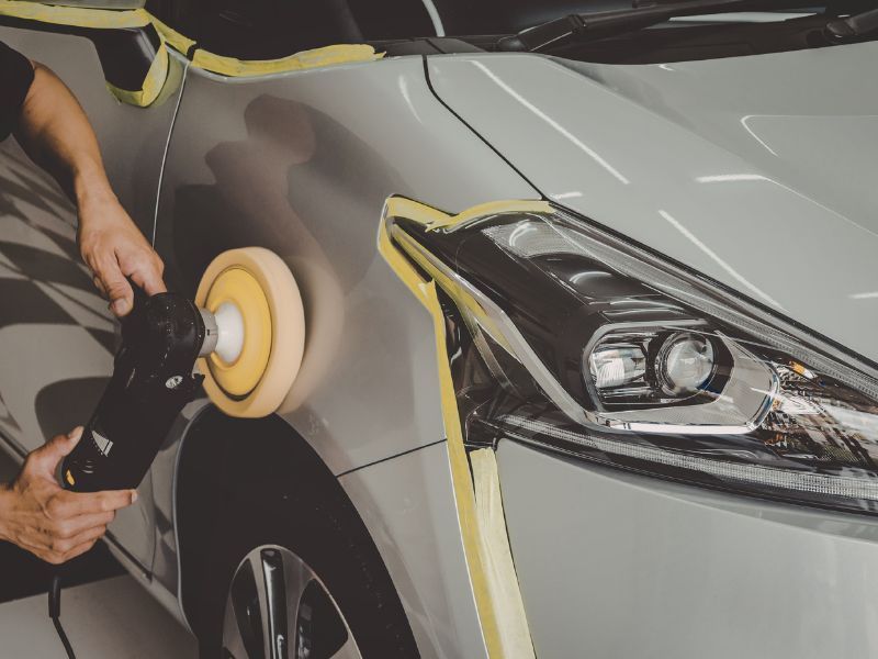 Person polishing a silver car with a buffer, masking tape around the headlight and fender