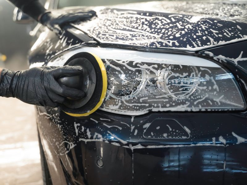 Gloved hand polishing a black car headlight with soapy foam on the hood