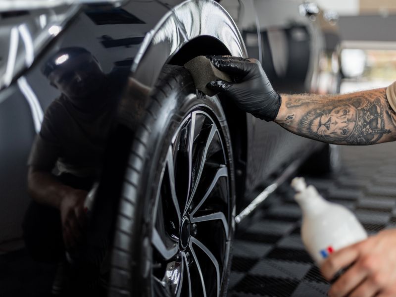 Person cleaning a black car wheel with a cloth and spray bottle in a garage
