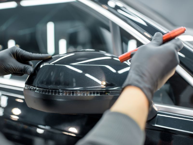 Gloved hands polishing a black car&rsquo;s side mirror with a red detailing tool