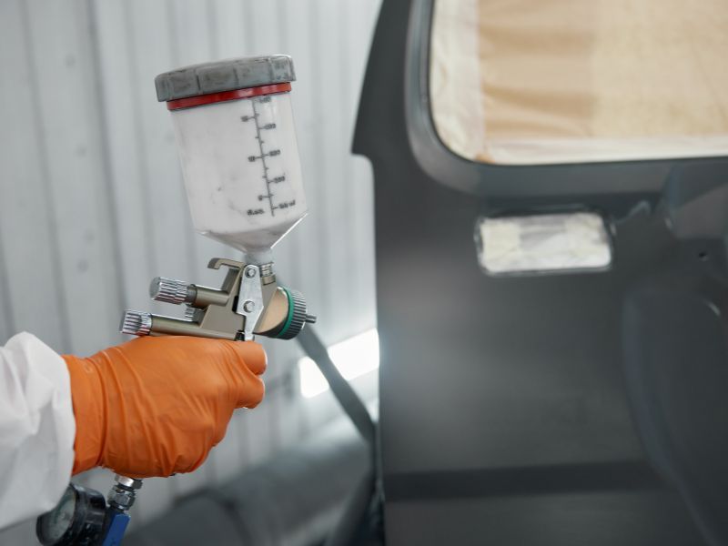 Gloved hand spraying paint with a spray gun near a car door in a workshop