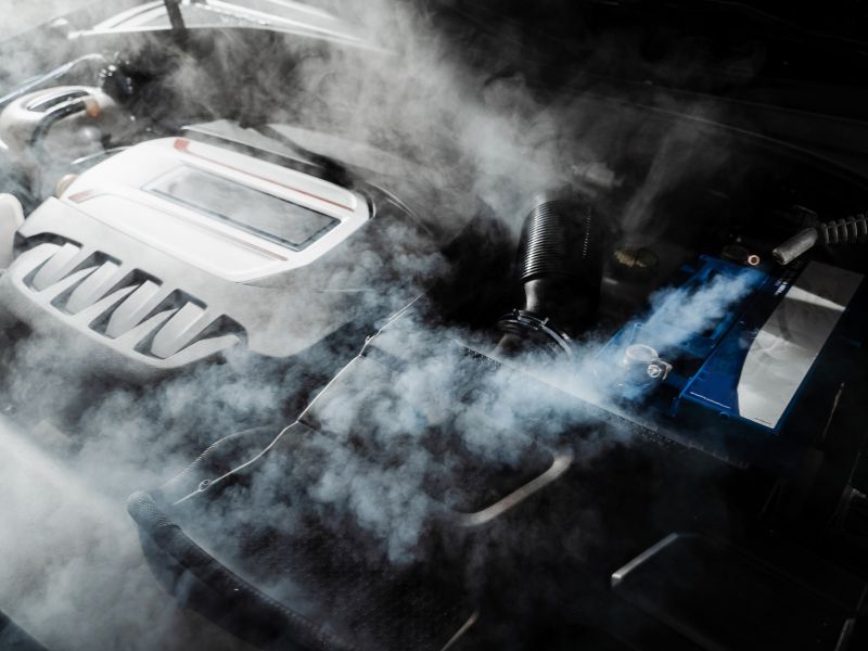 Smoky car engine bay with a vented engine cover and blue battery terminals under the hood