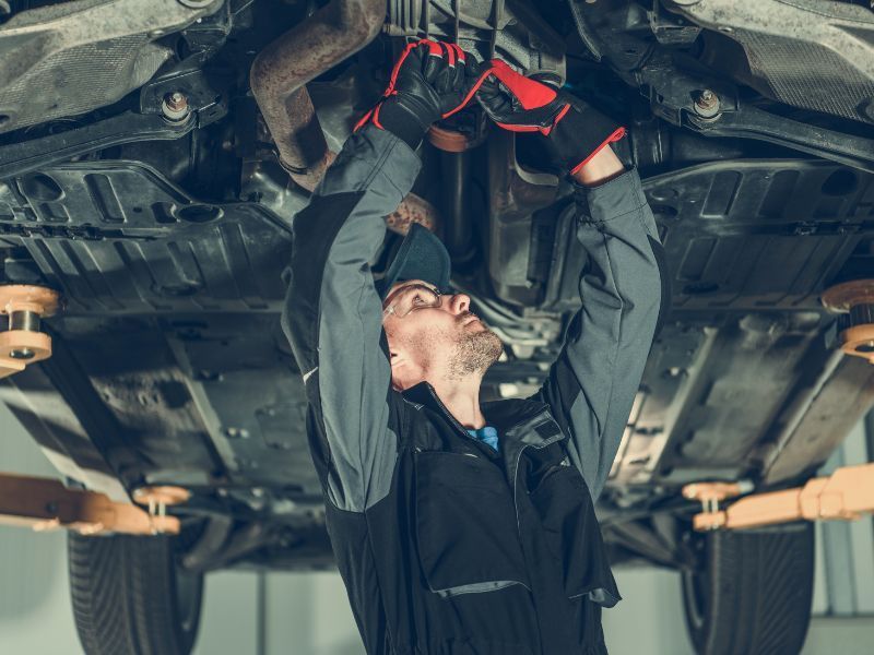 Mechanic working underneath a car on a lift, wearing gloves and looking up at the undercarriage