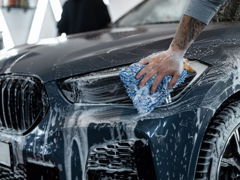 Person washing a black car with a blue sponge and soap suds on the hood