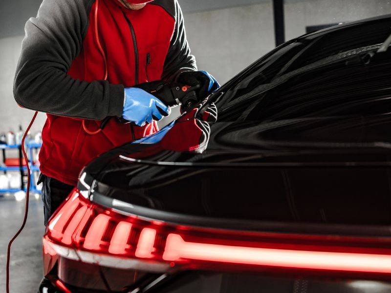 Mechanic in red vest polishing a black car’s rear with a buffer in an auto shop