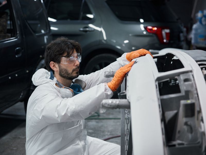 Worker in white coveralls and orange gloves inspects a car bumper in a garage