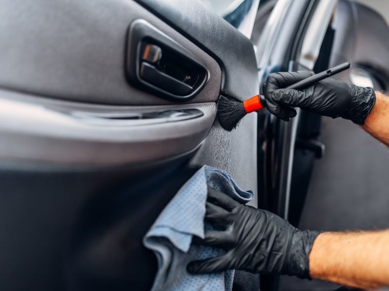 Gloved hands clean a car door interior with a cloth and detailing brush