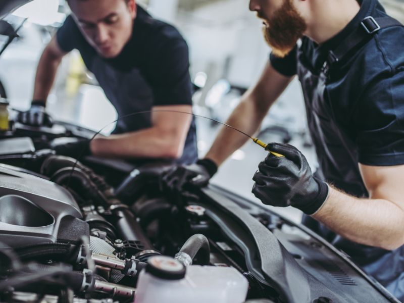 Two mechanics inspecting a car engine in a garage, one holding a tool and leaning over the hood