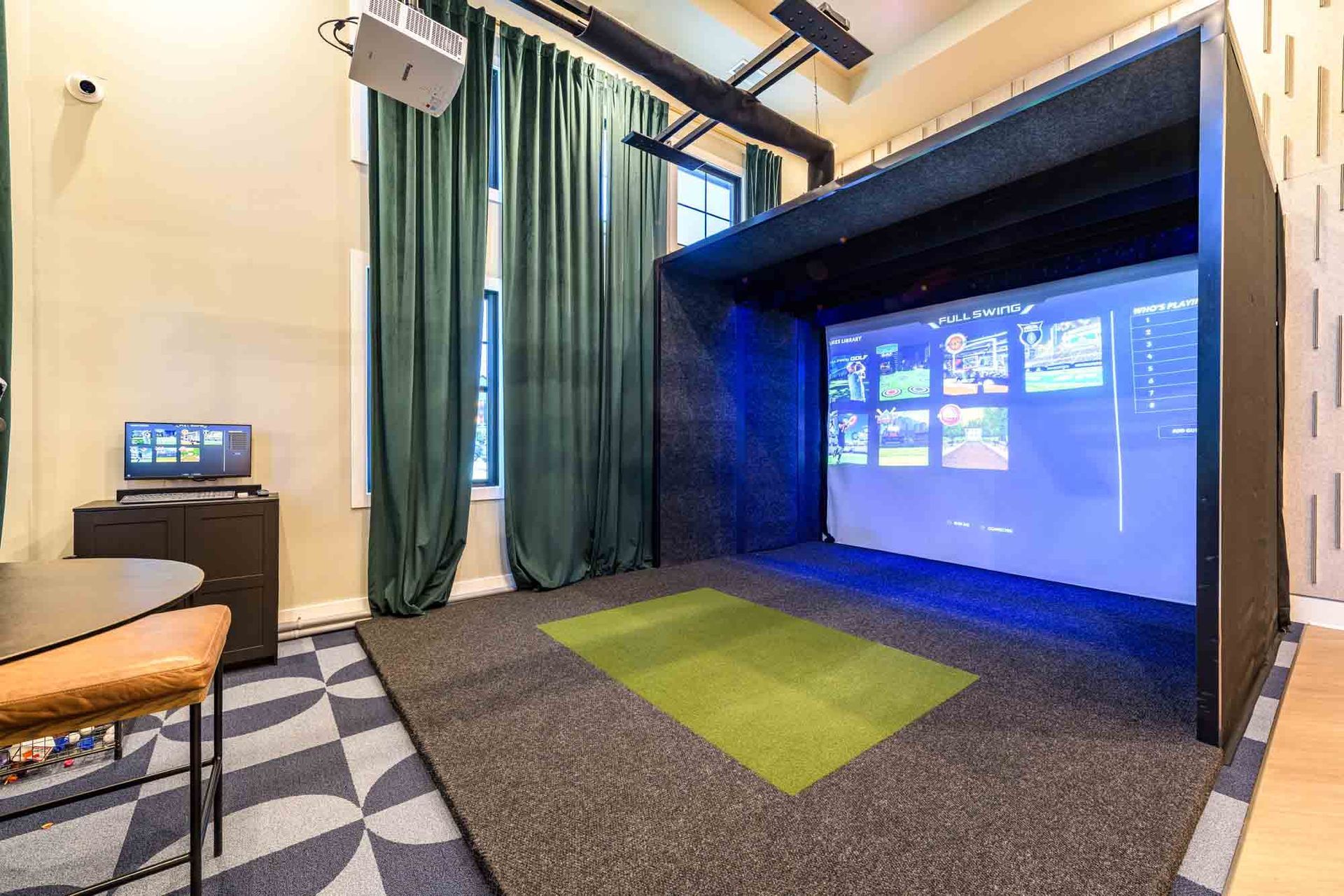 Golf simulator room with a screen displaying a user interface, black walls, green mat, and dark blue curtains.