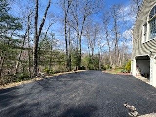 Black asphalt driveway leading into a wooded area with a house on the right. Blue sky.