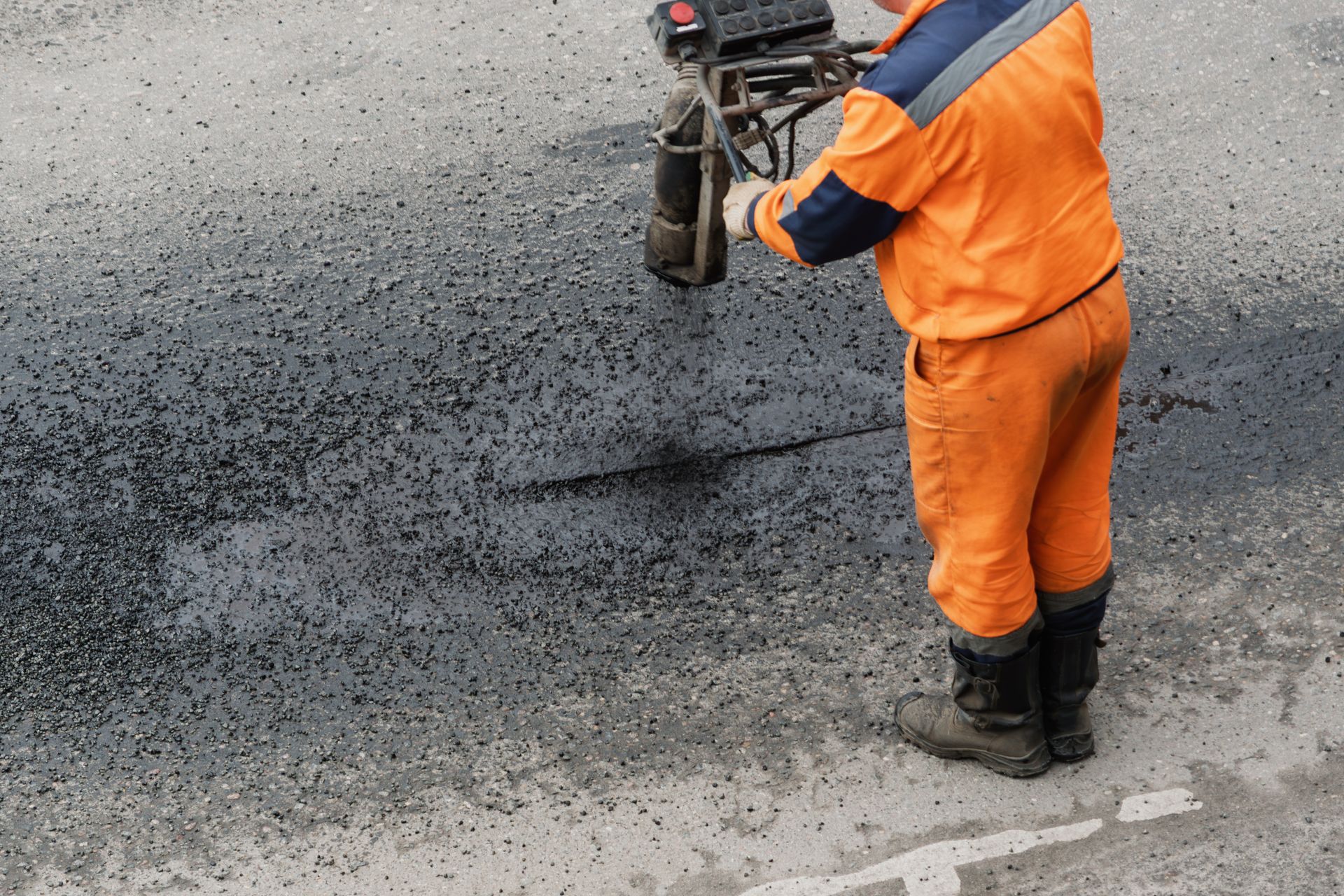 Road worker in orange safety gear using equipment to patch asphalt on a street.