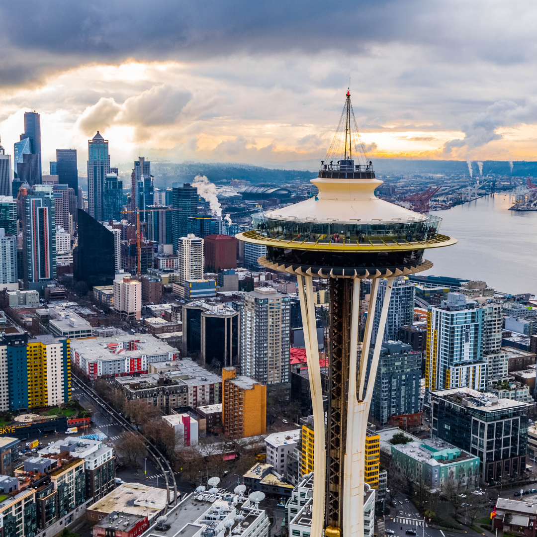 Seattle cityscape with the Space Needle in the foreground, skyscrapers, water, and cloudy sky.