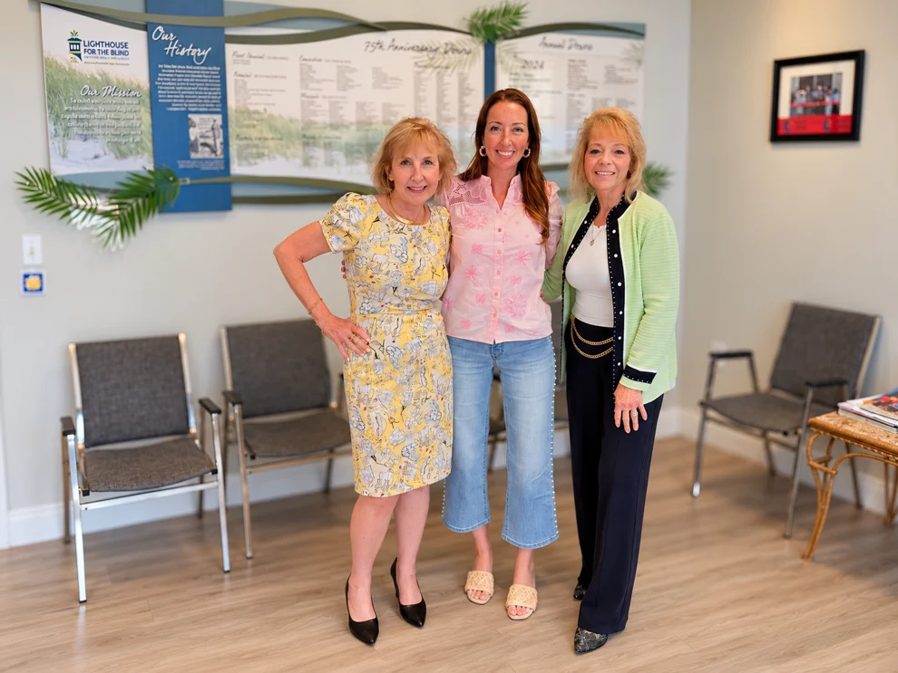 Three women stand together smiling in a Lighthouse for the Blind office, with the organization’s history display, chairs, and framed artwork in the background.