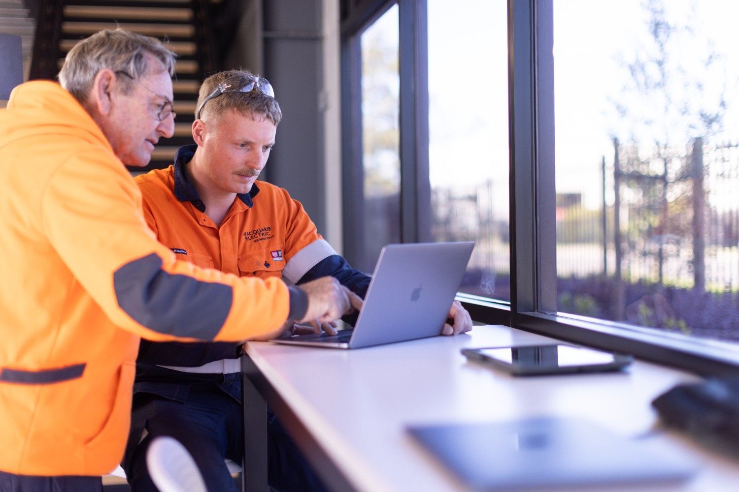 Two Men Are Sitting at A Table Looking at A Laptop Computer — Macquarie Electric in Newcastle, NSW