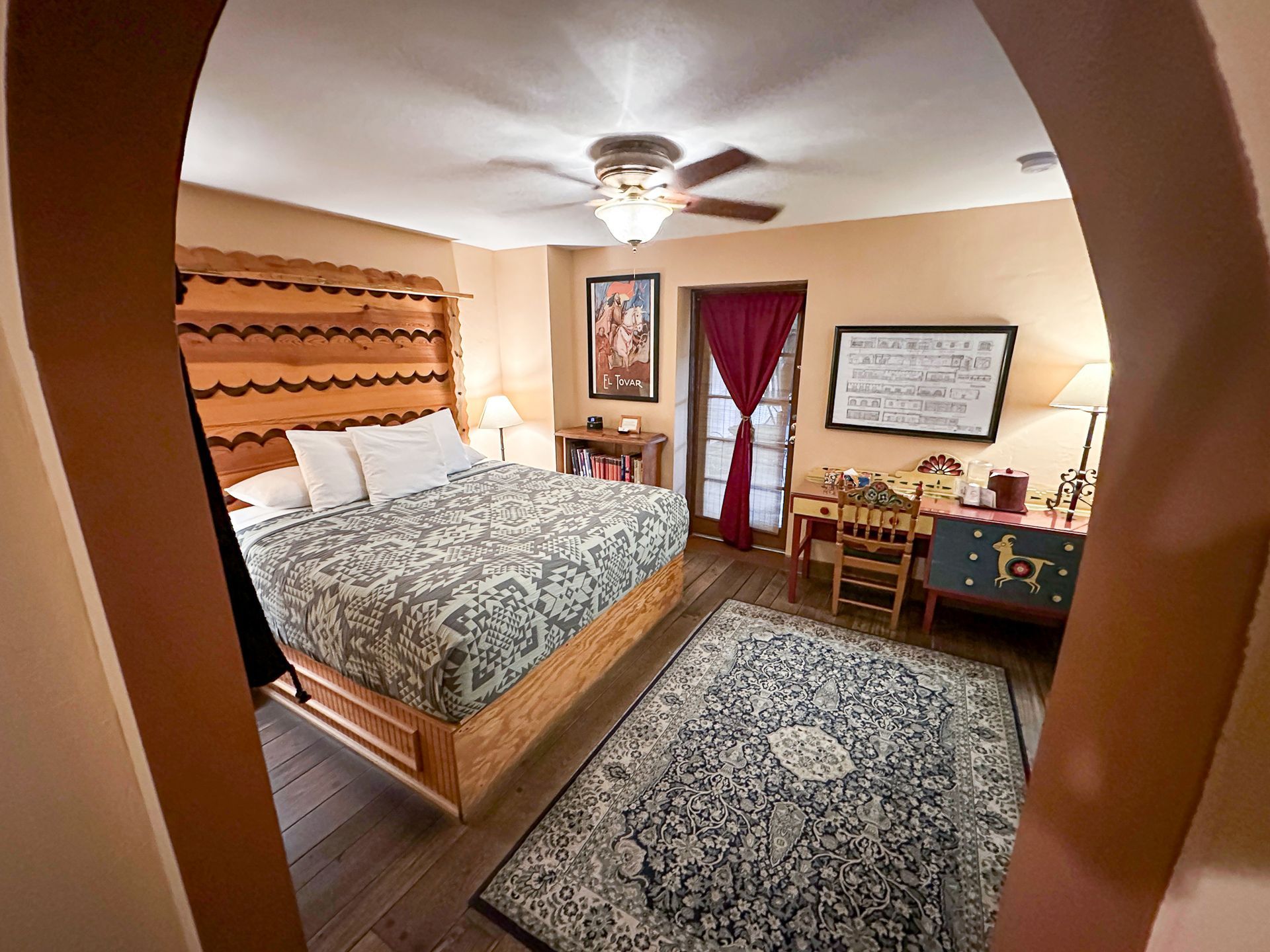Bedroom with bed, desk, rug, and arched doorway. The room has wood accents and warm colors.