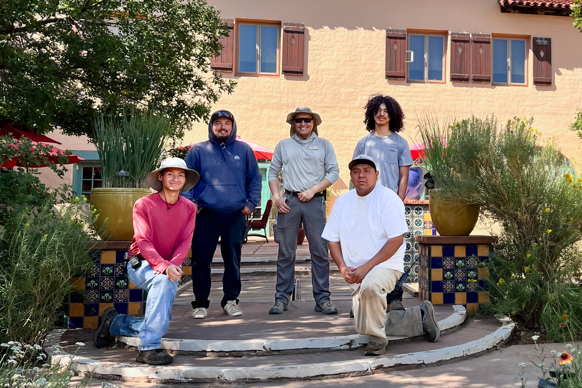 Five people posing on outdoor steps in front of a tan building with tiled planters and trees.