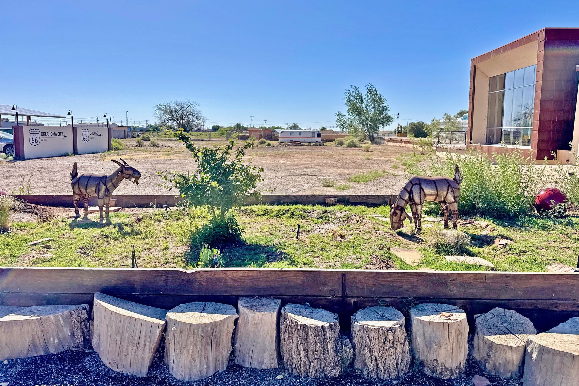 Two donkeys grazing in a dry field beside a modern building and stone border under a clear blue sky