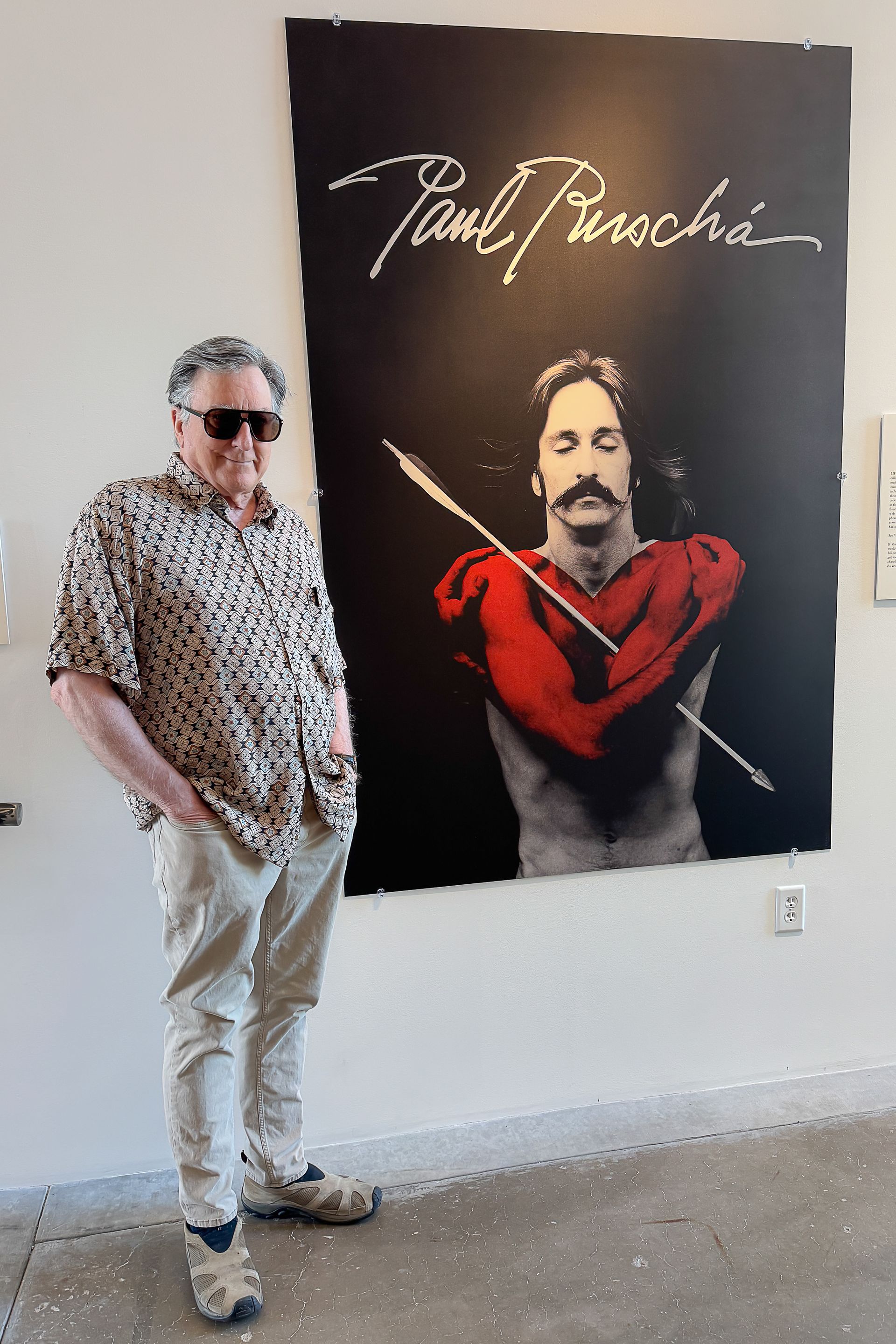 Man standing near a large poster with his name Paul Ruschá and a self portrait called Martyr for Love. 