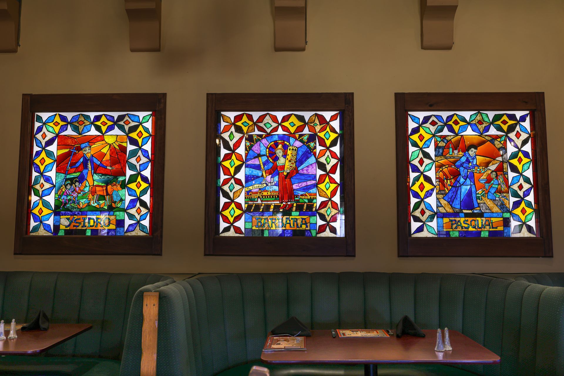 Three colorful stained-glass panels with figures hang on a wall above a green booth.