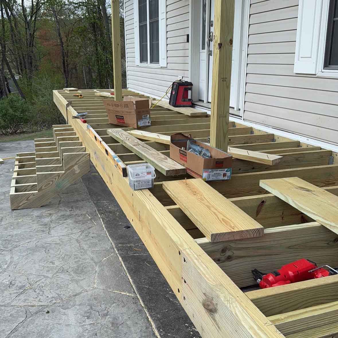 Deck construction in progress: wood frame, stairs, posts, tools, and boards near a house.
