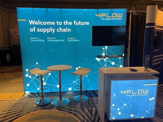 Trade show booth for 4flow, offering supply chain solutions. Teal and white backdrop with table, stools, and screen.