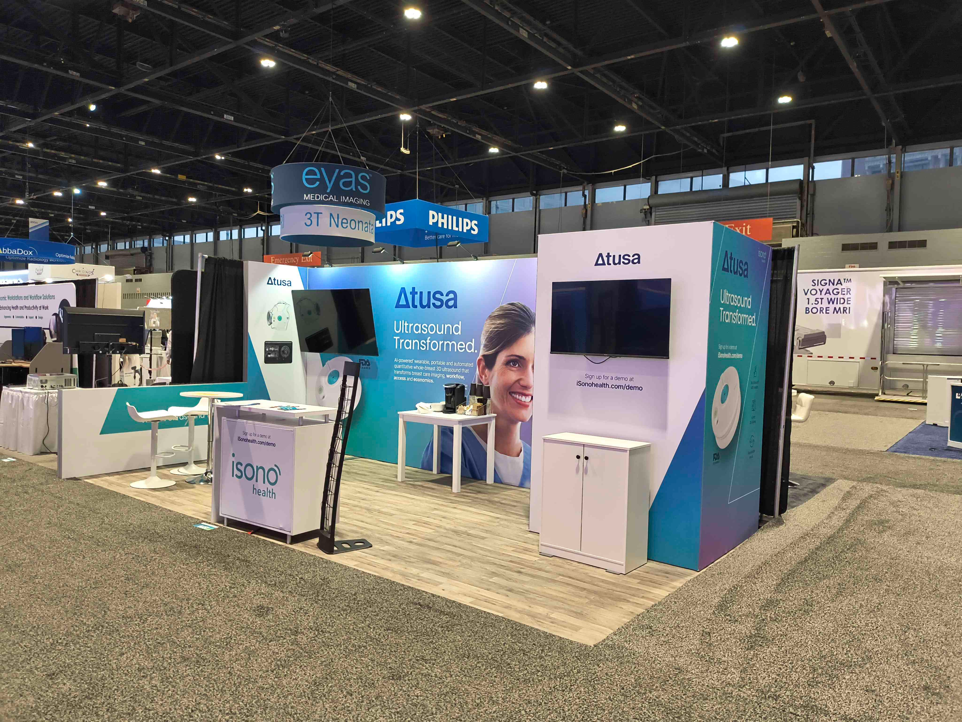 Trade show booth with white and teal accents, featuring screens and brand logos.