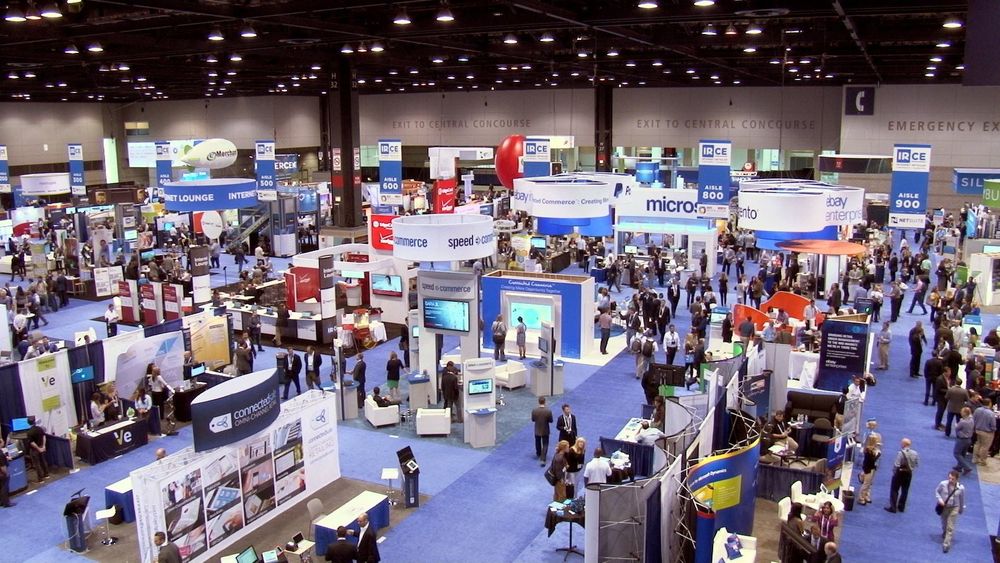 Trade show floor with booths, people, and product displays. Blue carpet and many colorful signs.