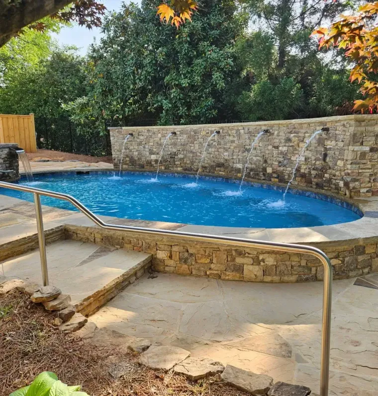 A stone-walled, oval backyard pool with a built-in water feature, surrounded by paved decking and lush trees.