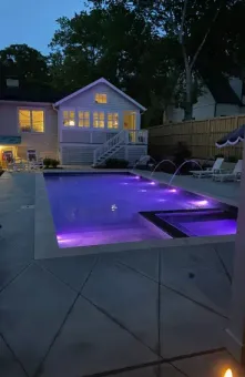 A backyard pool and attached spa illuminated in purple at dusk, with water jets, a patio, and a lit house in the background.