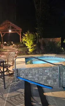 A backyard scene at night featuring an illuminated hot tub, an adjoining swimming pool, patio furniture, and a gazebo.