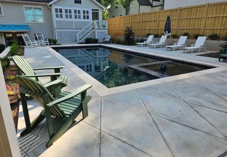 A rectangular backyard pool with a built-in spa, surrounded by a concrete patio, lounge chairs, and two green chairs.