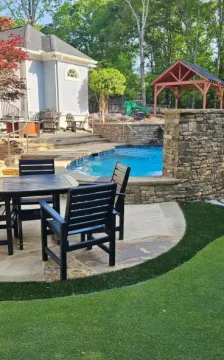 A backyard patio with a dining set on stone pavers, a small pool with a stone wall, and a putting green with a flag.
