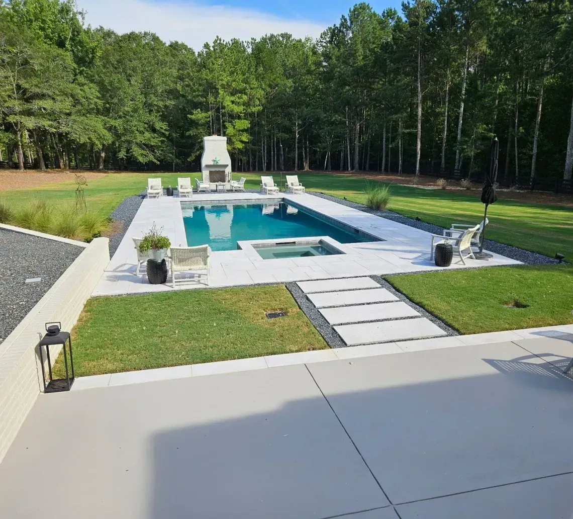 A rectangular swimming pool with a hot tub and stone fireplace set in a spacious backyard bordered by trees.