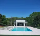 A rectangular swimming pool in front of a white pool house, surrounded by light stone decking and trees under a blue sky.