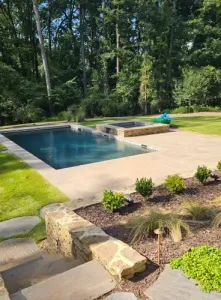 A backyard with a rectangular swimming pool, stone patio, stone steps, and landscaping next to a dense forest.