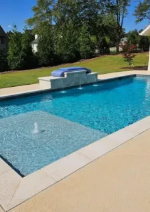 A rectangular backyard pool with a sun ledge, fountain, and stone coping, surrounded by a light-colored concrete deck.